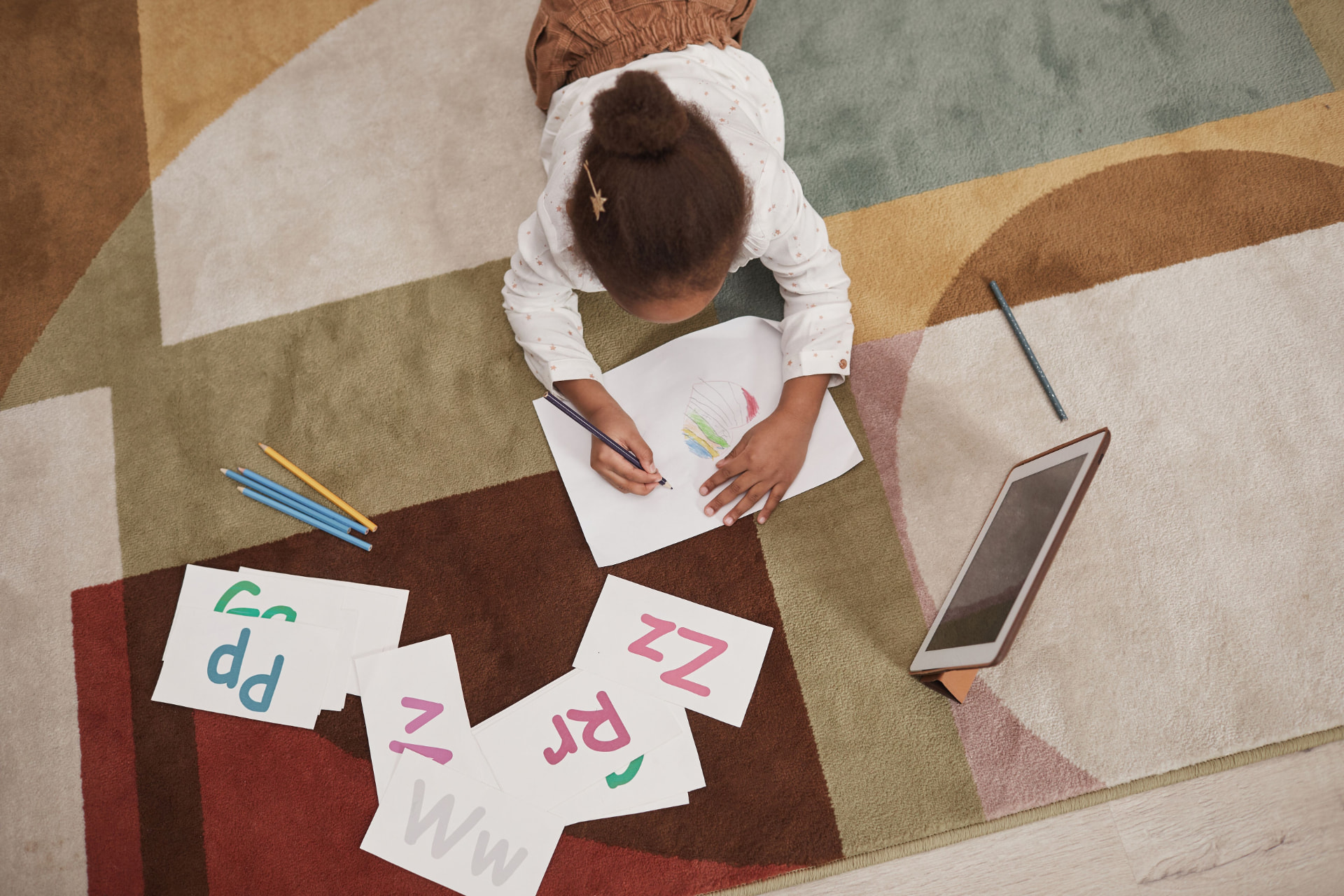 A young girl drawing on the floor, surrounded by colorful letter cards